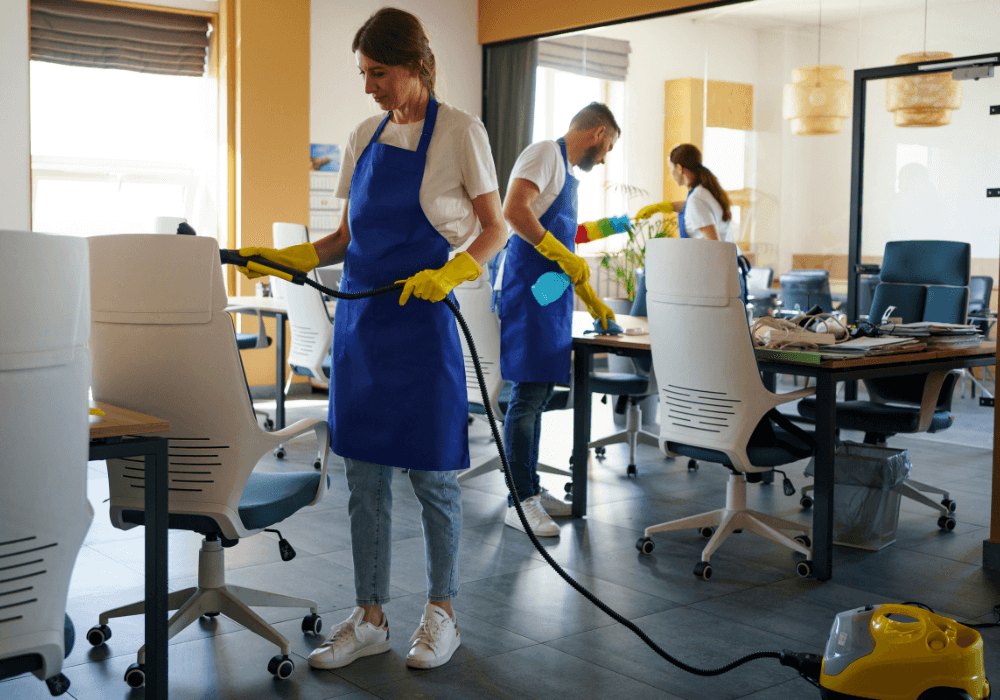 Three individuals in blue aprons and gloves clean an office space with a steam cleaner and cleaning supplies scattered around.