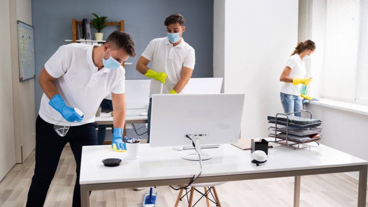 Three individuals cleaning an office space while wearing masks and gloves. One is wiping a desk, another is adjusting a monitor, and the third is cleaning a window.