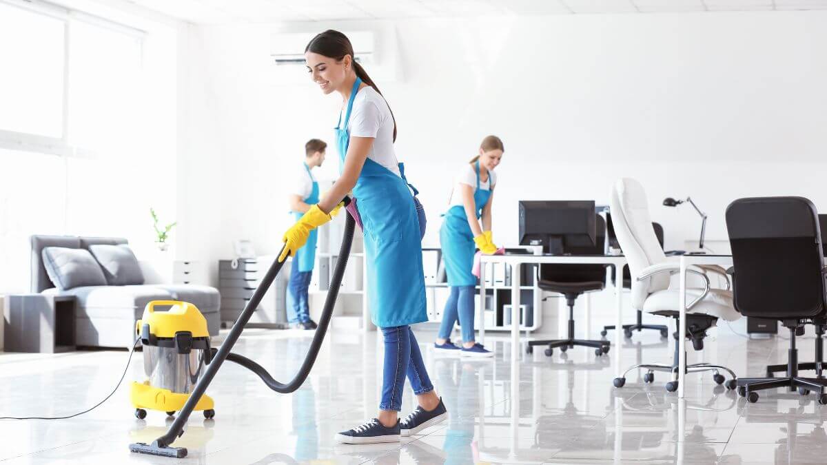 Three people, wearing blue uniforms and yellow gloves, are cleaning an office space using a vacuum cleaner and cloths. The room has a modern design with furniture and office equipment.