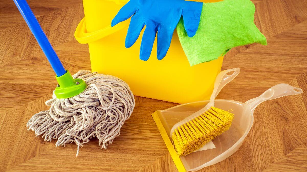 Cleaning supplies including a mop, yellow bucket, sponge, and dustpan with brush on a wooden floor.
