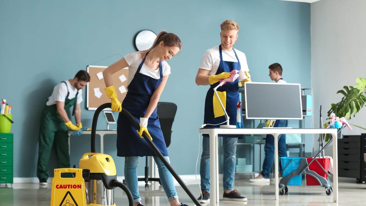 A group of people are cleaning an office space, using a vacuum cleaner, cleaning sprays, and other tools.